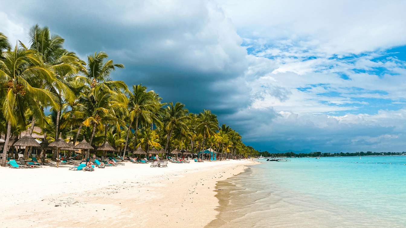 Plage de Trou aux Biches avec palmiers et huttes, eaux turquoise sous un ciel nuageux